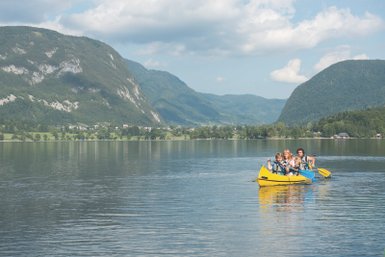 Eine Gruppe von vier Personen paddelt fröhlich in einem gelben Kajak auf dem ruhigen Bohinjsee, umgeben von grünen Bergen.