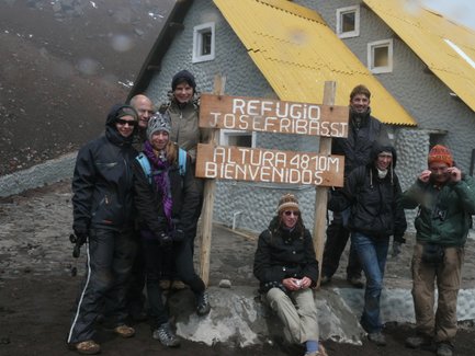 Eine Gruppe von sieben Personen posiert vor einem Refugium in den Bergen, umgeben von grauer Landschaft und einem nebligen Himmel.