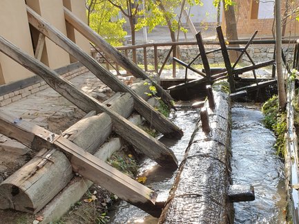 Ein Wasserlauf fließt über große, runde Baumstämme, die in einem alten Mühlenbereich angeordnet sind, umgeben von Bäumen.