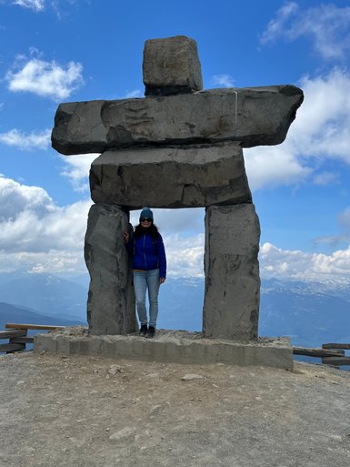 Eine Person steht in einem großen Steinskulptur, umgeben von einer beeindruckenden Berglandschaft und einem blauen Himmel.