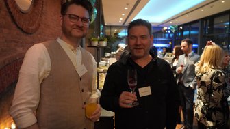 Zwei Männer stehen in einem eleganten Raum, einer hält ein Glas Sekt, der andere ein Getränk in der Hand.