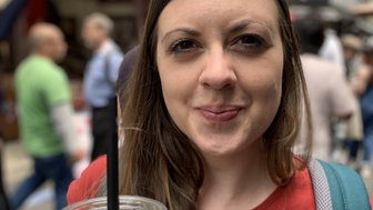 Eine Frau mit langen Haaren hält ein Getränk in einem belebten Markt, umgeben von Menschen und bunten Ständen.