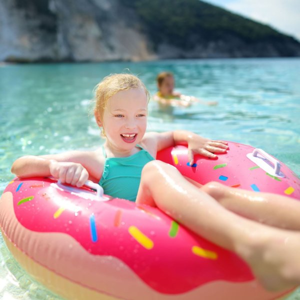 Ein fröhliches Mädchen mit blonden Locken genießt das Wasser in einem bunten Donut-Floatie an einem sonnigen Strand.