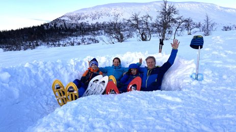 Eine Familie sitzt im Schnee, umgeben von schneebedeckten Bergen, und winkt fröhlich in die Kamera.