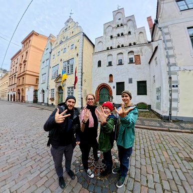 Vier Personen posieren fröhlich auf einer gepflasterten Straße in Riga, umgeben von historischen Gebäuden.