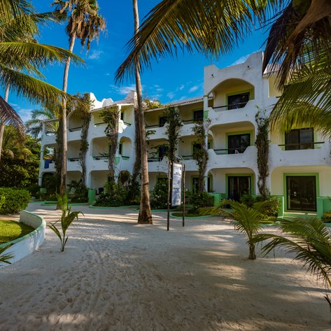 Ein helles, mehrstöckiges Gebäude mit grünen Akzenten und üppiger Vegetation umgeben von Palmen und feinem Sand.
