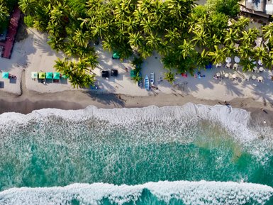 Eine luftige Perspektive zeigt den langen, ruhigen Strand mit palmengesäumter Küste und den sanft brechenden Wellen.