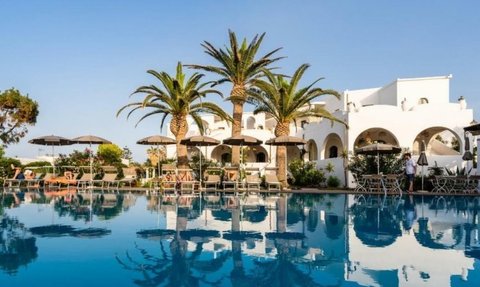 Ein luxuriöser Poolbereich mit Palmen und Sonnenliegen, umgeben von weißen Gebäuden unter klarem Himmel.