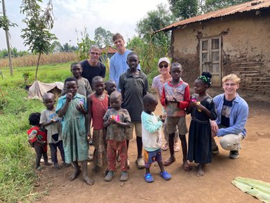 Eine Gruppe von Kindern steht fröhlich im Freien, umgeben von Natur und einem einfachen, rustikalen Hintergrund.