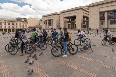 Eine Gruppe von Radfahrern steht auf einem Platz in Bogotá, umgeben von Tauben und historischen Gebäuden.