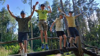 Vier Jungen balancieren fröhlich auf einem Baumstamm im Wald, umgeben von grünen Bäumen und strahlendem Himmel.