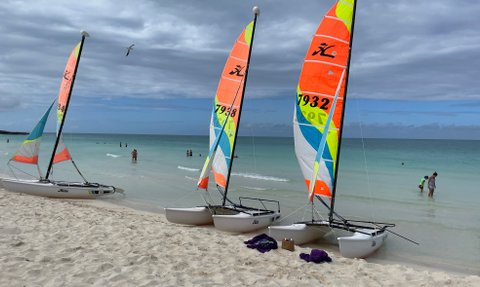 Drei bunte Katamarane stehen am Strand, während Menschen im klaren Wasser planschen und die Wolken am Himmel ziehen.