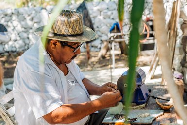 Ein älterer Mann mit einem Strohhut arbeitet konzentriert an einem Tisch, während er kleine Objekte in seinen Händen hält.