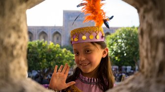 Ein fröhliches Mädchen mit einem bunten Hut und einer Feder winkt durch ein Fenster, umgeben von grünen Bäumen.