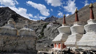 Drei weiße Stupas mit roten Spitzen stehen vor einer dramatischen Berglandschaft und einem Kloster im Hintergrund.