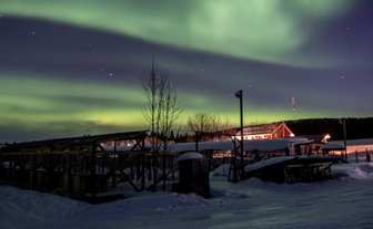 Ein atemberaubendes Nordlicht erhellt den Nachthimmel über einer verschneiten Landschaft mit Holzstrukturen im Vordergrund.