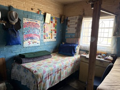 Ein einfaches Zimmer mit zwei Betten, bunten Decken und einer Fensterbank, die Tageslicht hereinlässt.