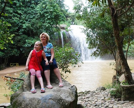Eine Frau und ein Mädchen sitzen auf einem großen Stein, umgeben von üppigem Grün und einem Wasserfall im Hintergrund.