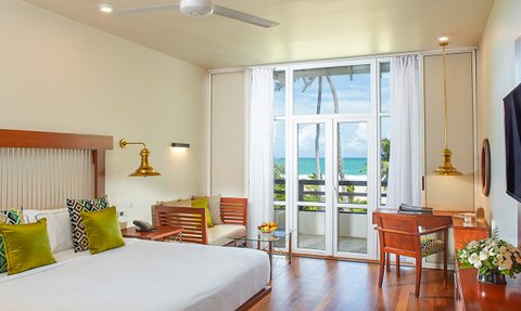 Ein helles Hotelzimmer mit einem großen Bett, bunten Kissen und Blick auf das Meer durch große Fenster.