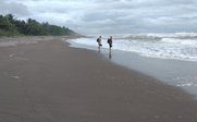 Zwei Personen stehen am Strand, während die Wellen sanft an den dunklen Sand schlagen, unter einem bewölkten Himmel.