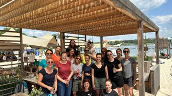 Eine Gruppe von Frauen posiert fröhlich unter einem Holzüberstand am Strand, umgeben von Wasser und Sonnenschein.