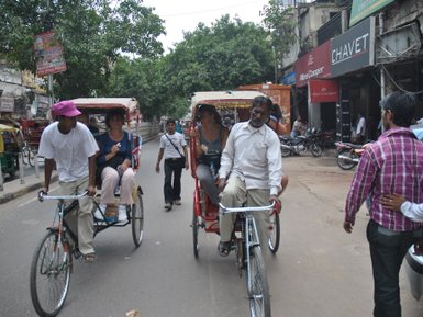 Zwei Rikschafahrer transportieren Touristen auf einer belebten Straße in Delhi, umgeben von Geschäften und Bäumen.
