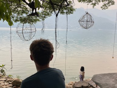 Ein Junge sitzt am Ufer des Phewa-Sees und beobachtet ein Mädchen, das am Wasser spielt, umgeben von grünen Bäumen.