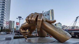 Eine große, goldene Statue einer Hand, die ein Schild mit der Aufschrift 'Gangnam Style' hält, steht in einer urbanen Umgebung.