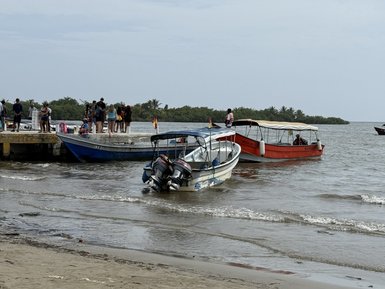Drei Boote liegen am Ufer, während Menschen auf einem Steg stehen und die ruhige Wasseroberfläche betrachten.