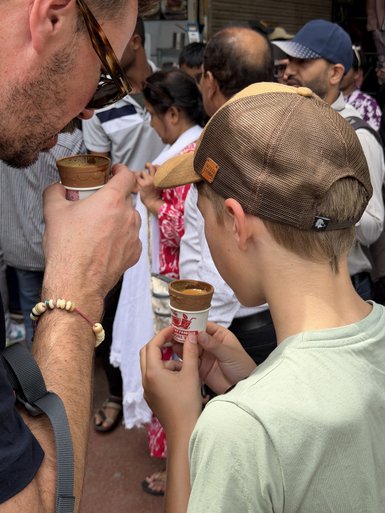 Ein Erwachsener und ein Kind genießen Chai-Tee aus kleinen Bechern, umgeben von einer lebhaften Menschenmenge.