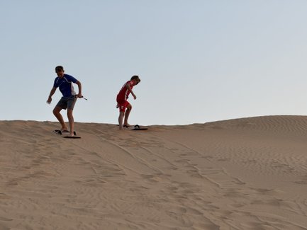Zwei junge Männer gleiten auf Sandboards die sanften Dünen einer Wüste hinunter, umgeben von goldenem Sand und klarem Himmel.