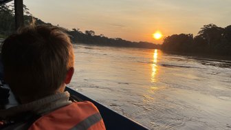 Ein Junge in einem orangefarbenen Schwimmwesten blickt auf den Sonnenuntergang über einem ruhigen Fluss in der Amazonasregion.