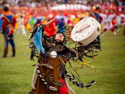 Ein junger Schamane tanzt leidenschaftlich auf einer Wiese, während er ein Trommelspiel begleitet und bunte Bänder schwingt.