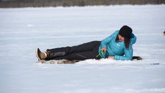 Eine Person liegt auf einer Schneedecke und hält ein grünes Werkzeug in der Hand, umgeben von einer winterlichen Landschaft.