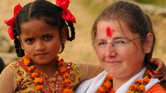 Eine Frau mit Brille lächelt, während sie ein einheimisches Mädchen mit Zöpfen und roten Schleifen umarmt.