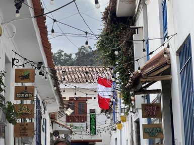 Eine enge Gasse in Cusco, Peru, mit weißen Wänden, bunten Schildern und einer peruanischen Flagge, die im Wind weht.