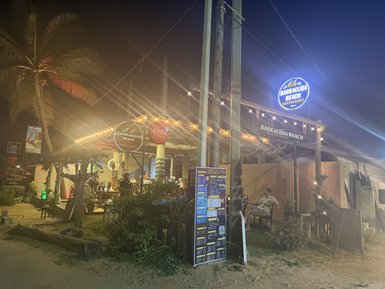 Ein einladendes Restaurant mit Lichtern und einem großen Schild, das den Namen 'Barracuda Beach' zeigt, umgeben von Palmen.