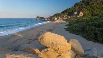Ein ruhiger Strand mit sanften Wellen, umgeben von großen Felsen und üppigem Grün im Hintergrund, unter klarem Himmel.