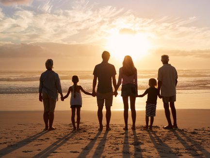 Eine mehrgenerationale Familie steht am Strand und beobachtet den Sonnenuntergang, während die Wellen sanft ans Ufer rollen.