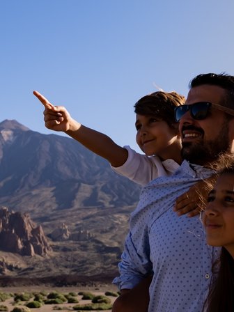 Eine Familie steht vor einer beeindruckenden Berglandschaft und zeigt auf etwas im Hintergrund, während sie lächeln.