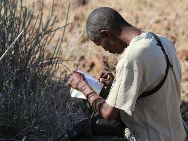 Ein Mann sitzt im Freien und notiert etwas in einem Notizbuch, umgeben von trockener Vegetation.