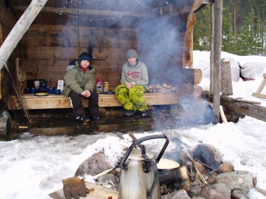 Zwei Kinder sitzen auf einer Holzbank in einer Hütte, umgeben von Schnee und Rauch vom Lagerfeuer.