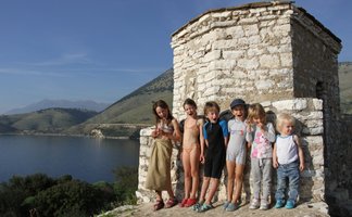Eine Gruppe von fröhlichen Kindern steht an einem steinernen Gebäude am Wasser, umgeben von grünen Hügeln und blauen Himmel.