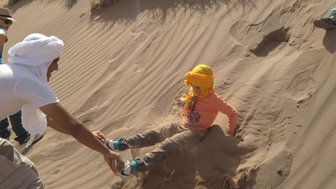 Ein Kind sitzt im Sand einer Düne, während ein Erwachsener ihm hilft, sich herauszuziehen, beide lachen fröhlich.