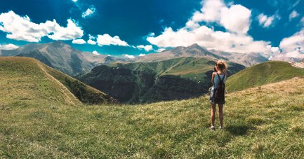 Eine Person steht auf einer grünen Wiese und blickt auf majestätische Berge unter einem strahlend blauen Himmel.