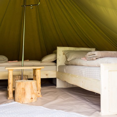 Im Inneren eines Zeltes stehen zwei Holzbetten mit gestreiften Bettlaken und gefalteten Decken, umgeben von einem warmen, grünen Zelt.
