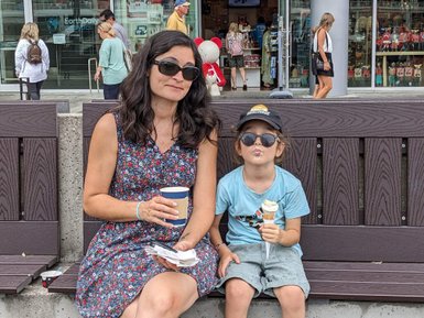 Eine Frau in einem blumigen Kleid sitzt auf einer Bank und genießt einen Kaffee, während ein Kind neben ihr ein Eis isst.