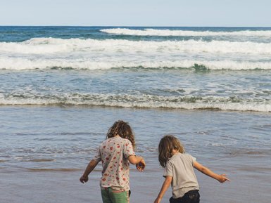 Zwei Kinder mit lockigen Haaren stehen im flachen Wasser am Strand und genießen den Moment, während die Wellen sanft an den Strand rollen.
