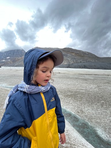 Ein Kind in einem blauen und gelben Regenjacke steht auf einem Gletscher, umgeben von grauen Wolken und Bergen.