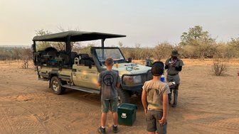 Ein Jeep steht in der Wüste, während zwei Kinder und ein Guide sich um einen Tisch mit Snacks versammeln.